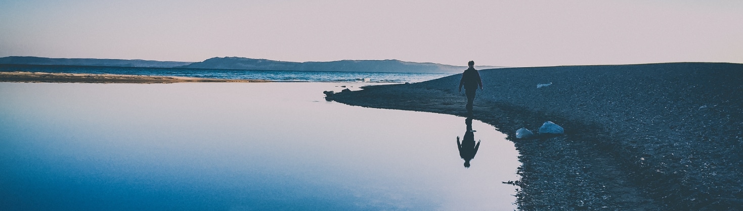 A man walking on a rocky beach at sunset near a calm lake.
